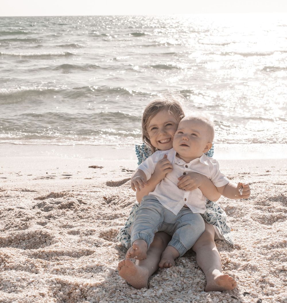 Two children play and laugh together while sitting on a sandy beach near the ocean.