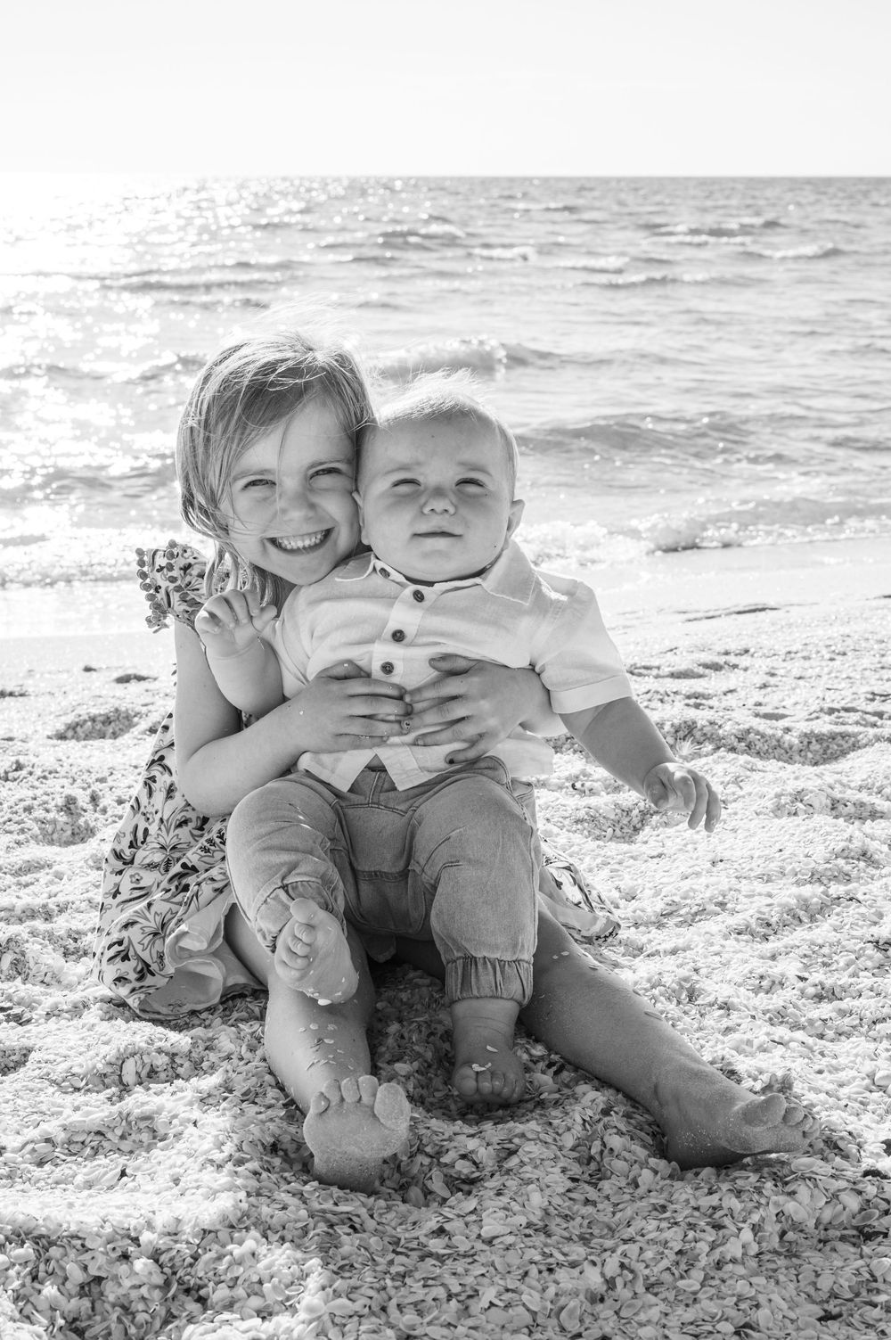 A black and white beach photo showing two children sitting together on the sand with ocean waves in the background.