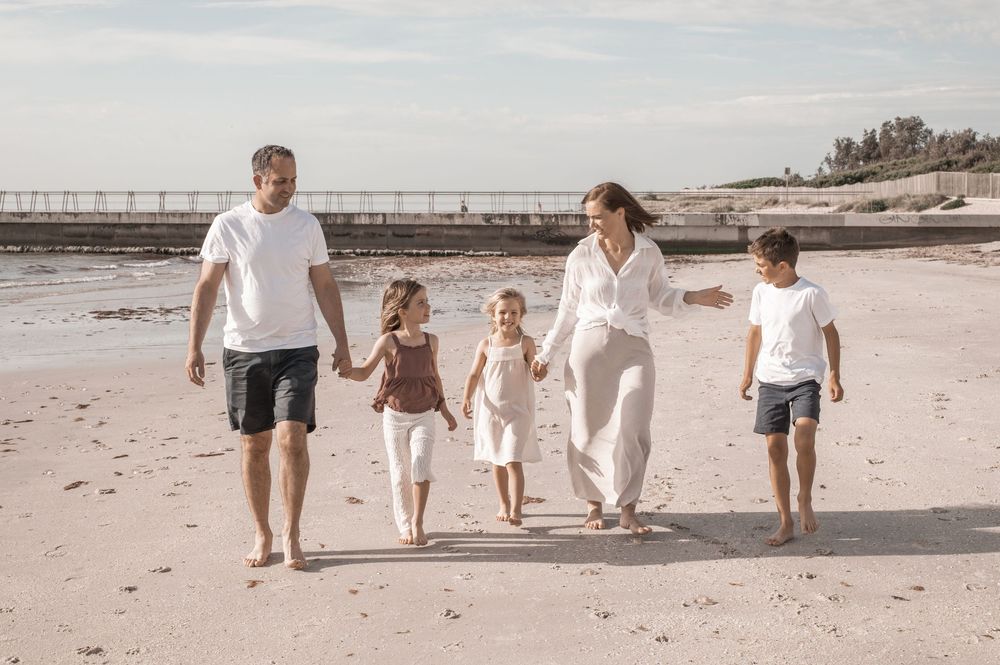 A family of five walking together barefoot on a sandy beach during a sunny day.