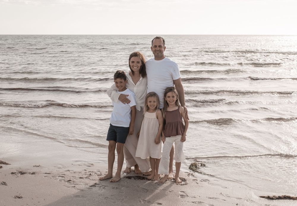 A family in coordinating neutral outfits walks along a sandy beach at sunset with waves rolling in.