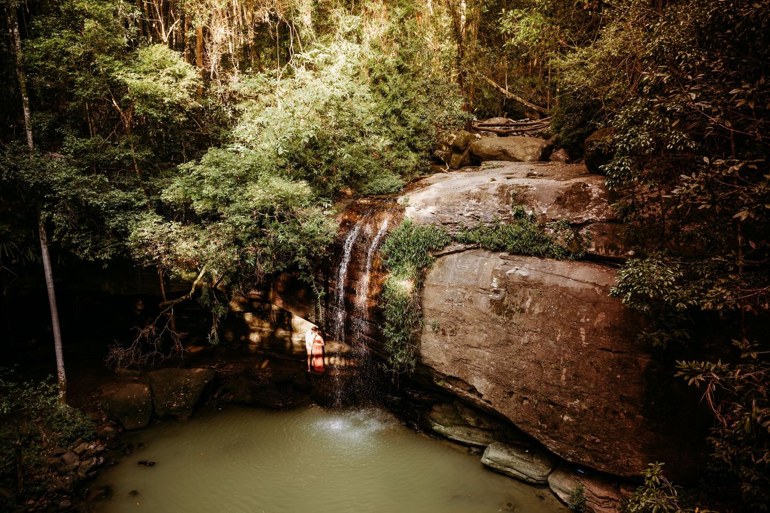 Person in red dress exploring a scenic waterfall grotto with rock formations and natural pool.
