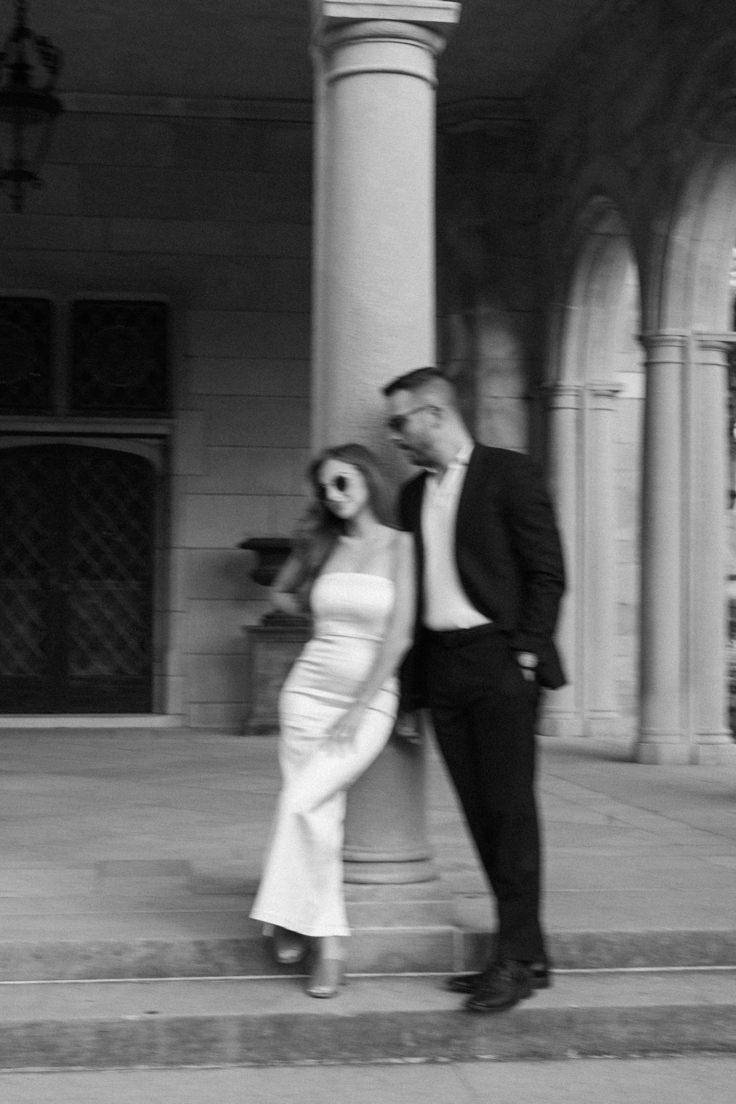 Stylish couple posing in black and white beside a stone column at a classic architectural venue.