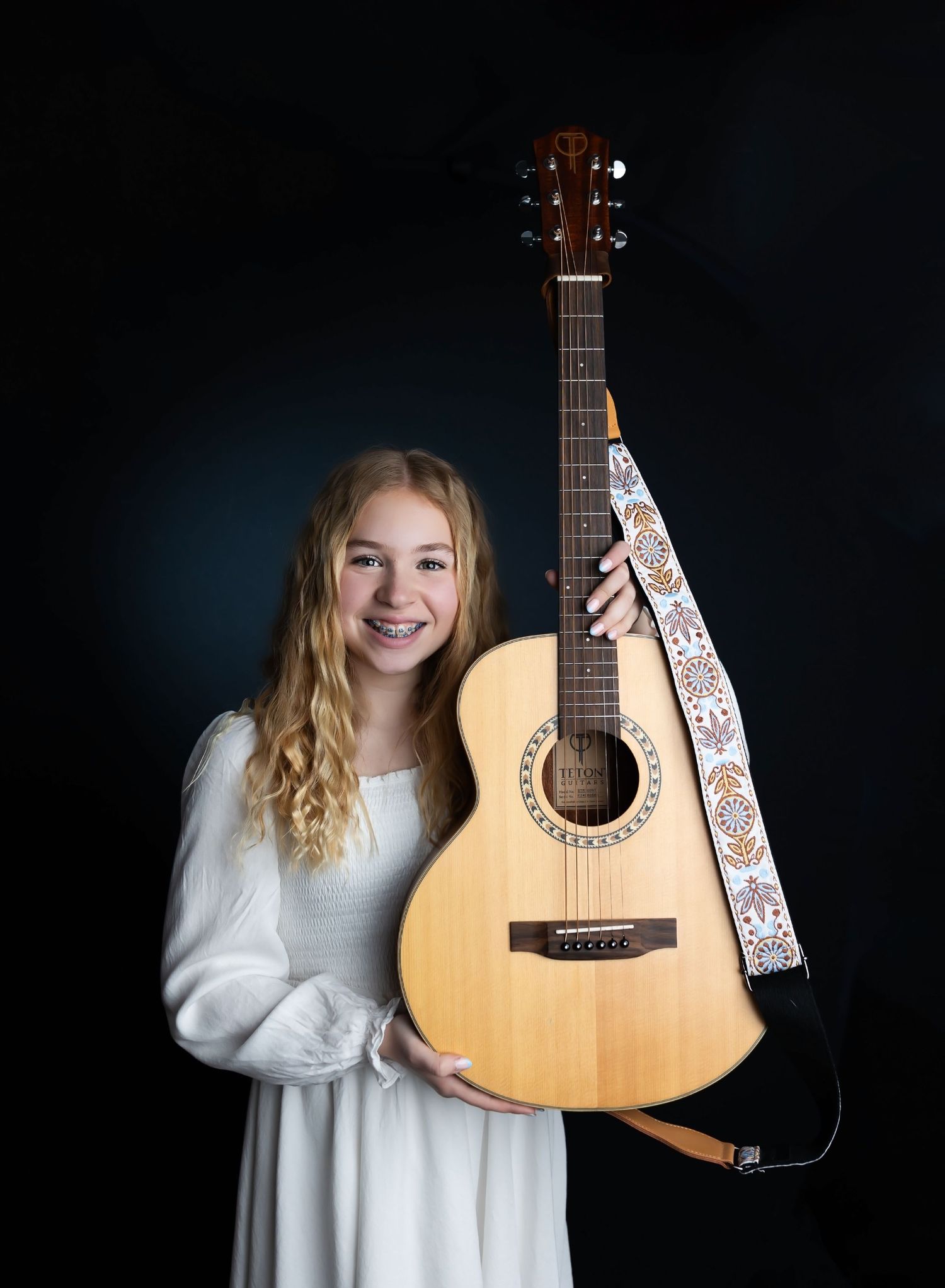 A musician in a white sweater dress with an acoustic guitar against a dark background in a photo series.
