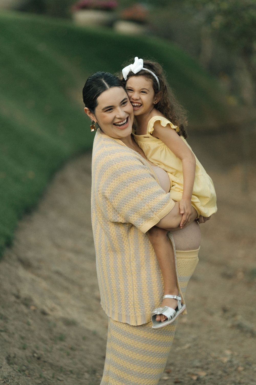 A laughing woman in a yellow striped outfit holds a smiling little girl in a yellow dress outdoors.