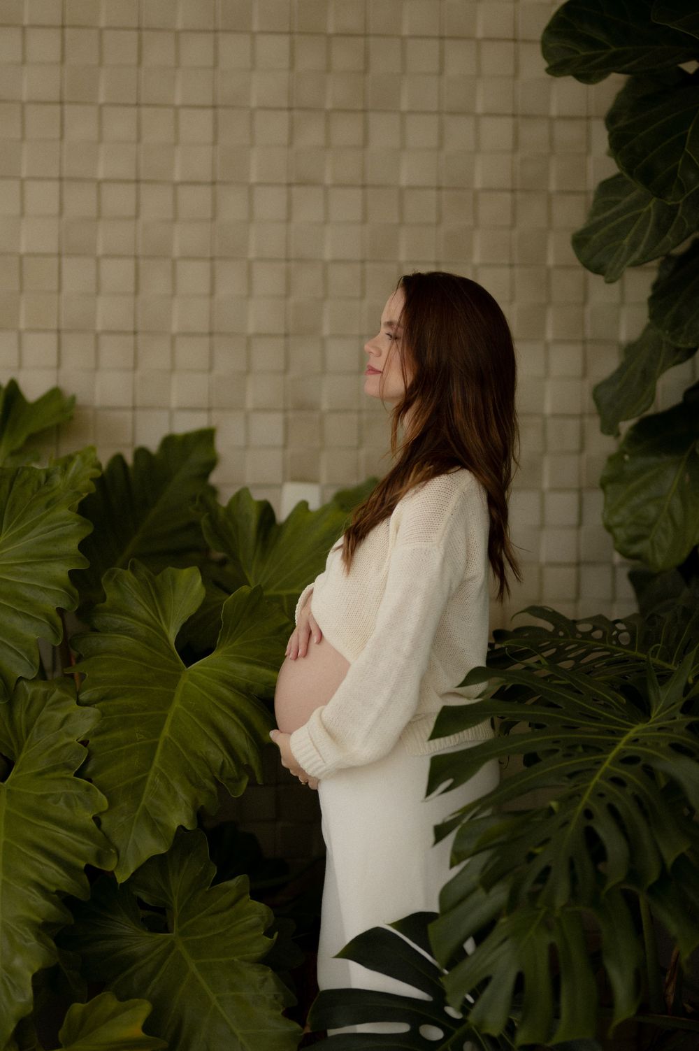 Pregnant woman in white dress stands among tropical green leaves touching her baby bump.