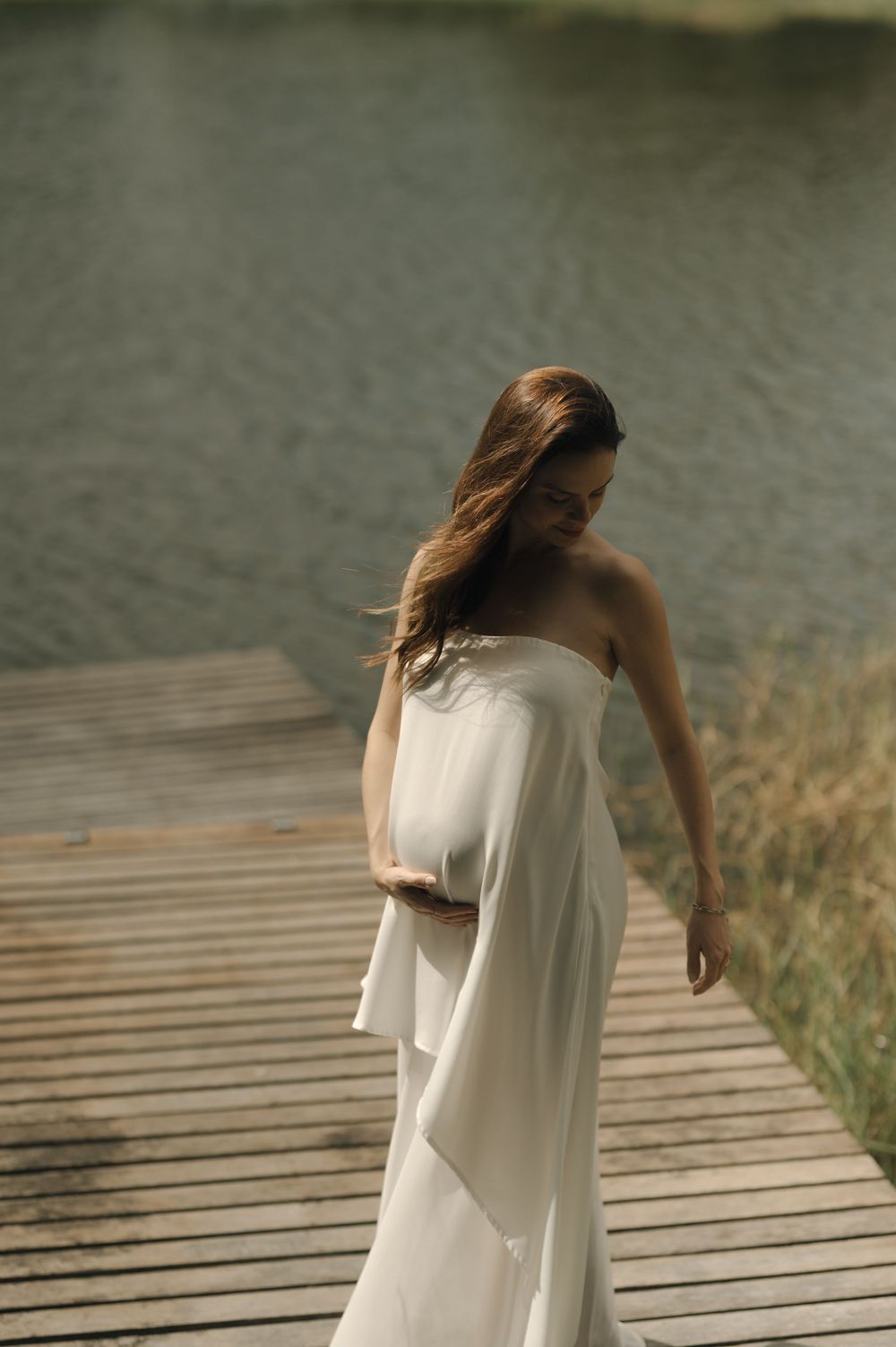 A woman in a flowing white strapless gown stands on a wooden dock by a serene lake, her hair gently blowing.