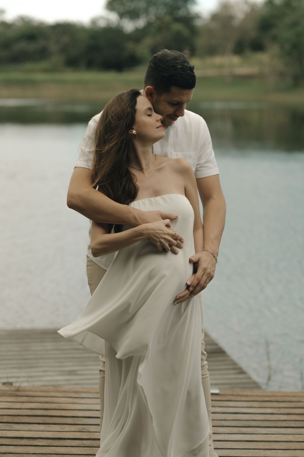 A couple shares a tender embrace on a wooden dock by a lake, the woman wearing an elegant white strapless gown.