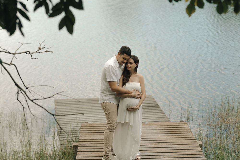Couple embracing on wooden dock by serene lake, woman in white maternity dress, man in white shirt, lush greenery.