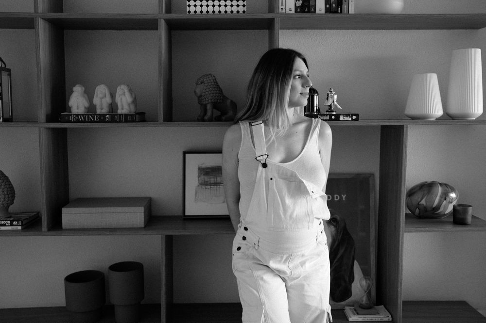 Woman in white outfit standing by modern shelving unit with decor, black and white photo.