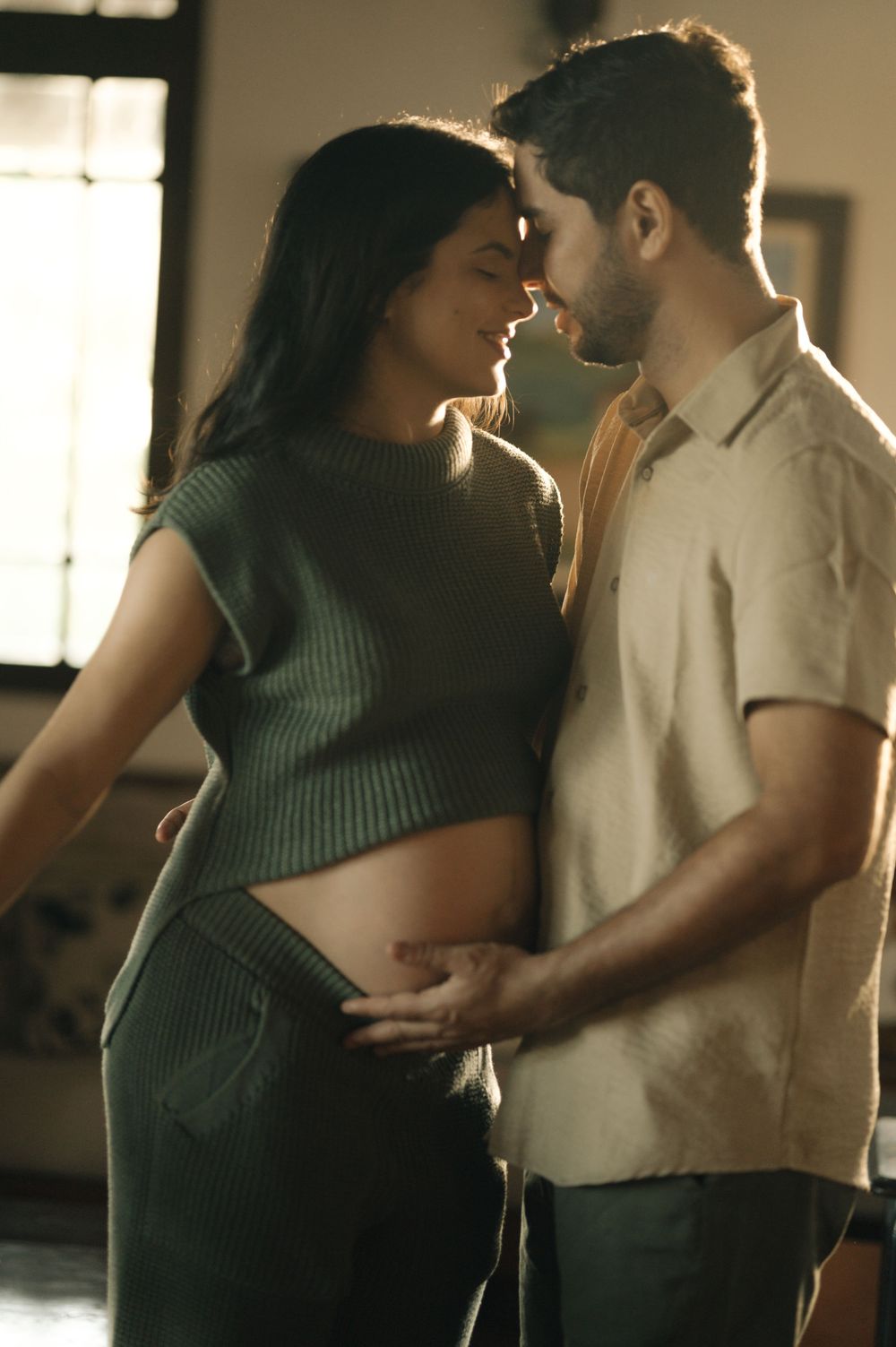 Couple sharing tender moment, man gently touching pregnant woman's belly in warm indoor setting.