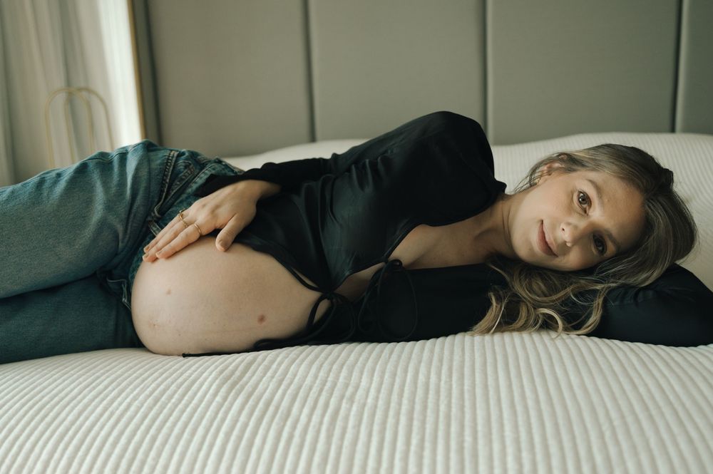 Pregnant woman lying on bed in black top and jeans, smiling and cradling baby bump during maternity photo shoot.