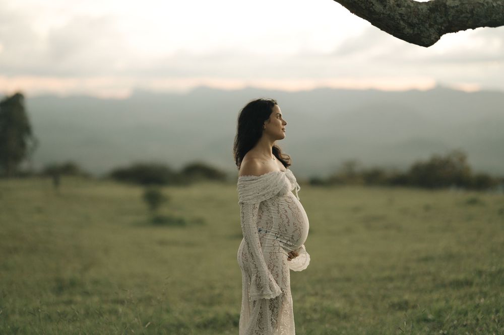 Pregnant woman in white lace dress cradling baby bump outdoors in green field with misty mountains backdrop.