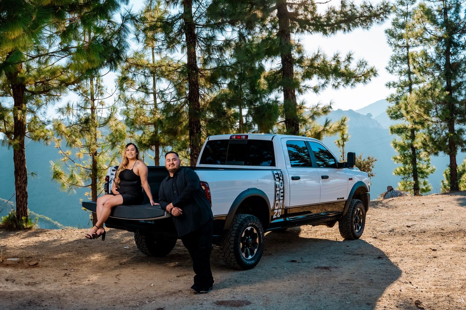 Two people sitting on the tailgate of a white pickup truck with mountain scenery behind them.