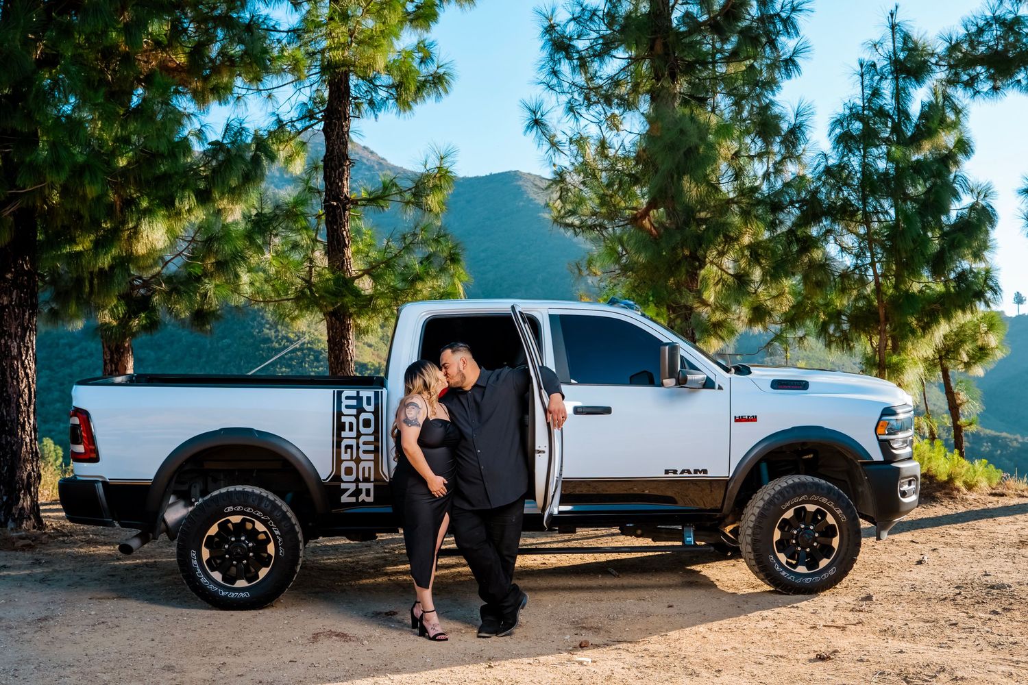A couple poses beside a lifted white pickup truck on a mountain overlook.