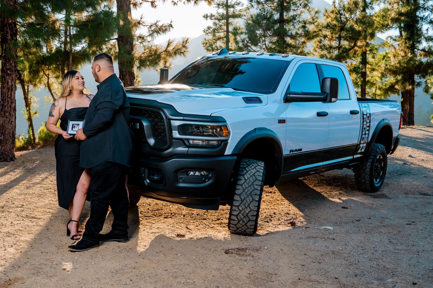 A couple poses against their lifted white Ram truck with mountains in the background.