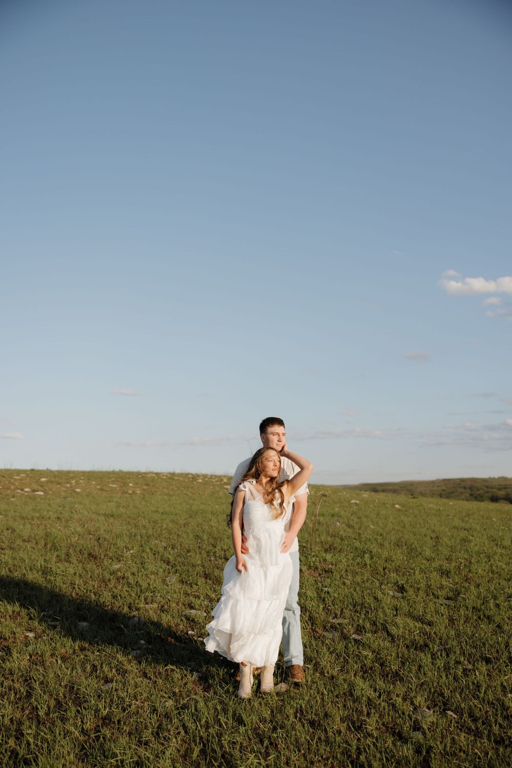 Couple stands in open green field under blue sky.