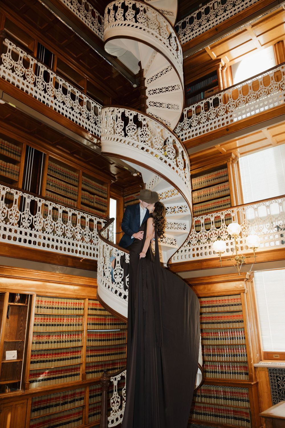 A spiral staircase with ornate white metalwork leads up through a library with wooden bookshelves lining the walls.