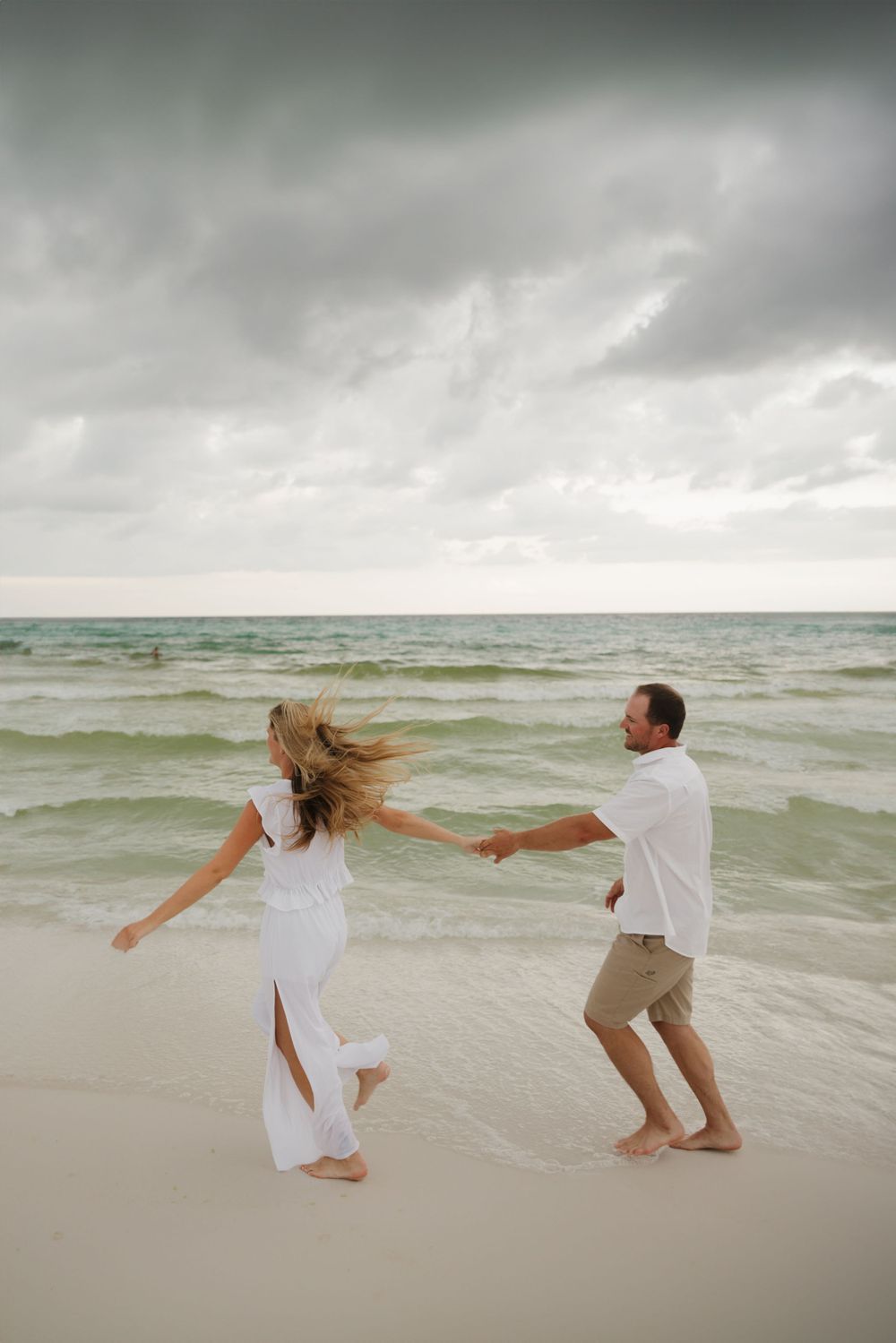 A couple in white clothing dances playfully on a stormy beach.