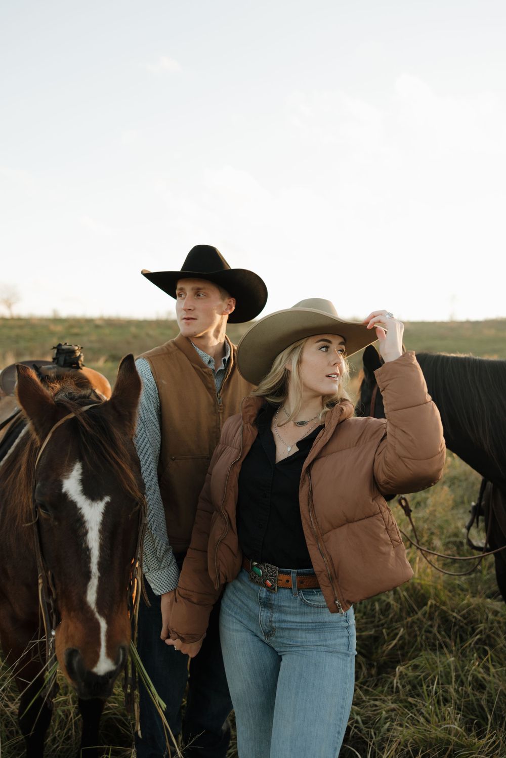 Western-styled couple pose with horses in rustic field setting.