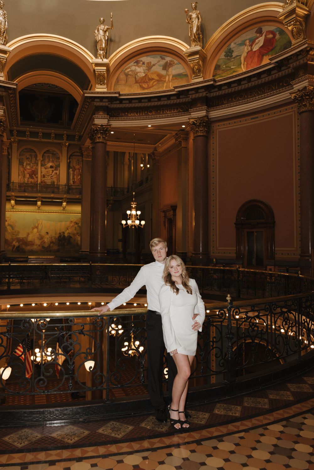 A couple stands together on an elegant balcony with ornate classical murals and architecture.