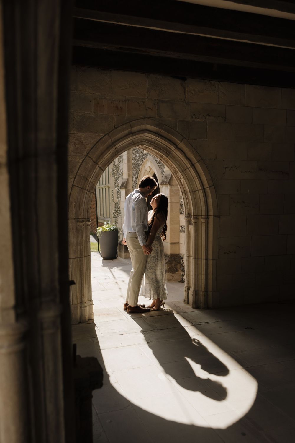 A silhouetted couple shares a romantic moment under a gothic stone archway as sunlight creates dramatic shadows on the floor.