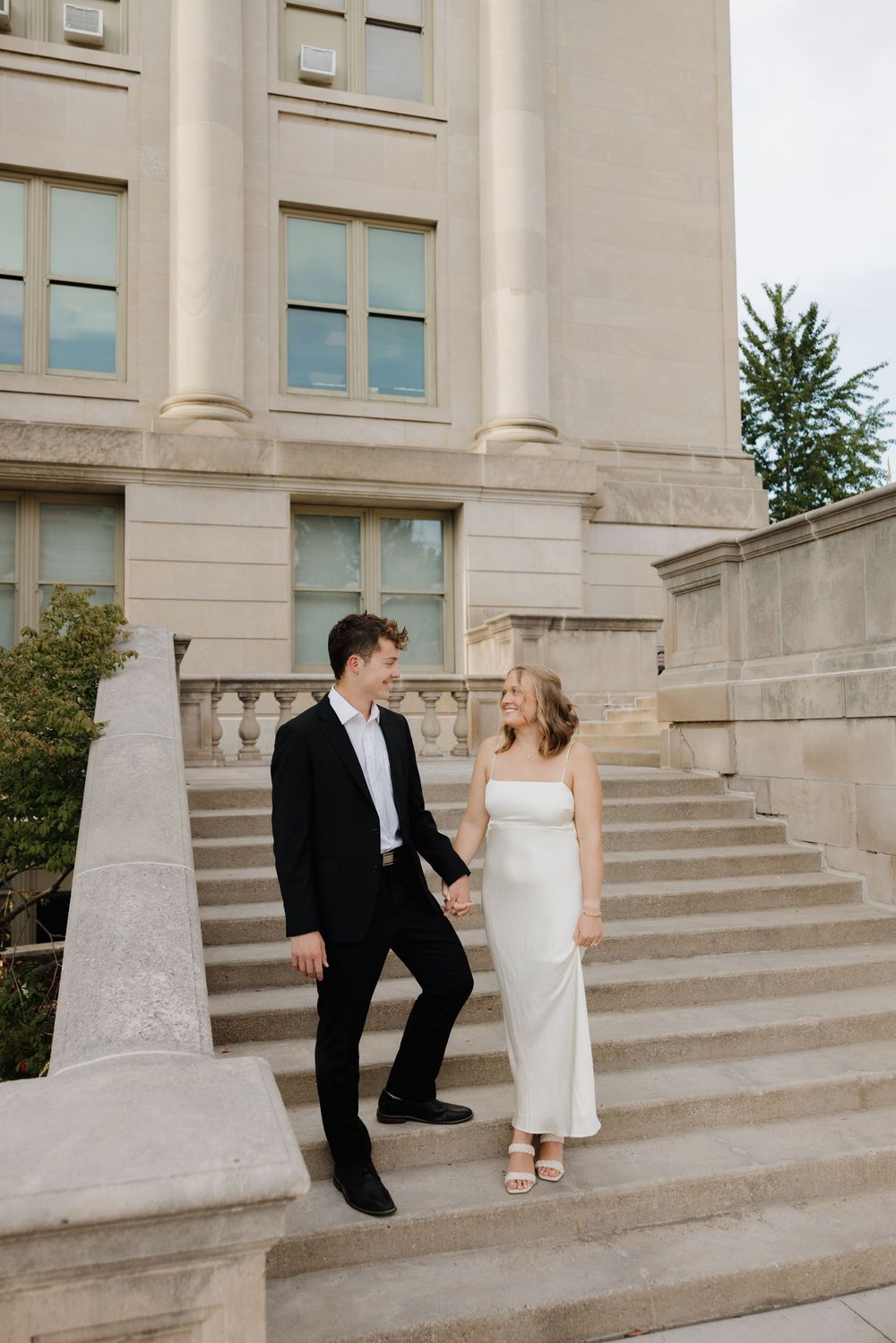 A wedding couple shares a playful moment sitting on stone steps outside a grand building.
