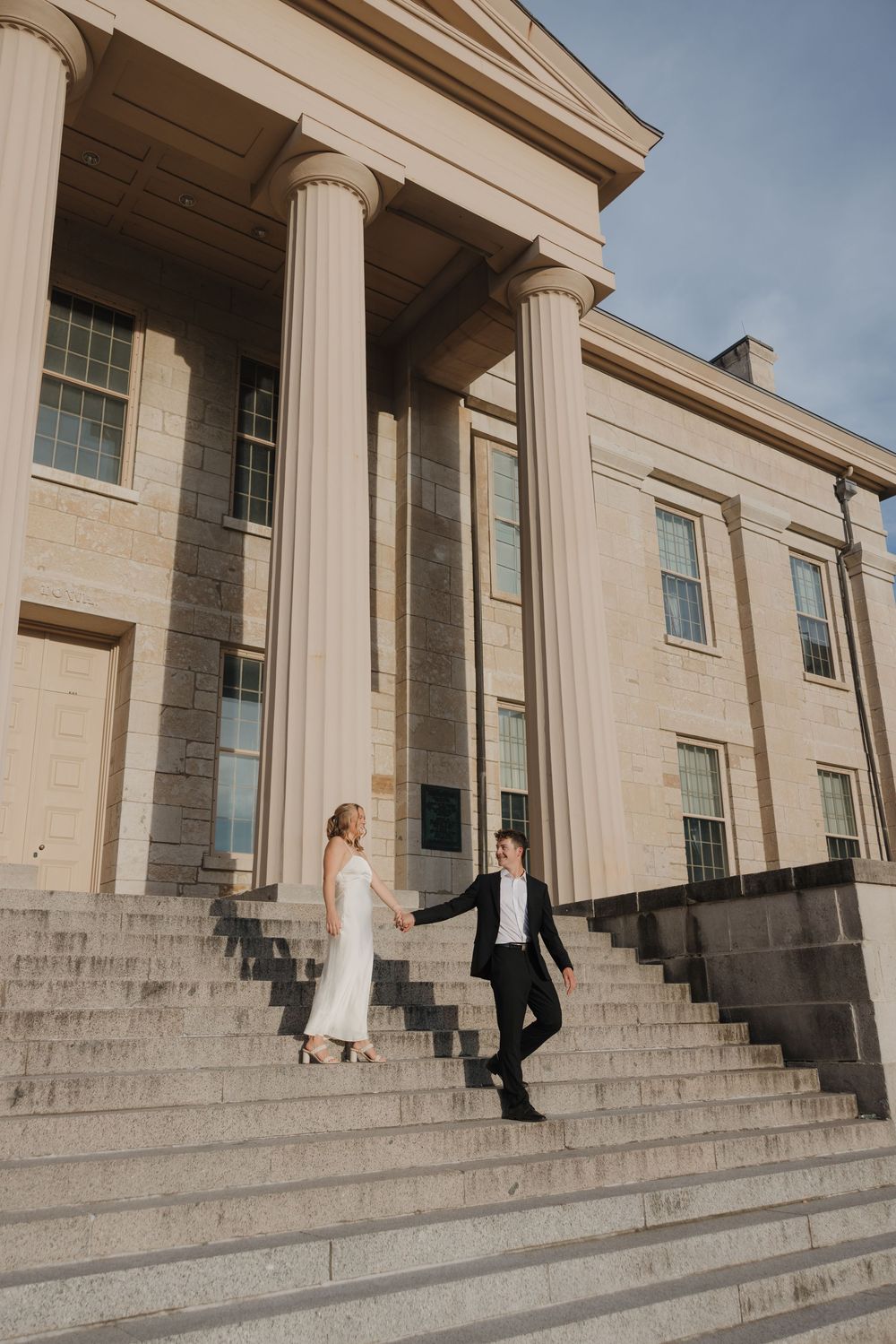 A couple walks down grand stone steps of a neoclassical building with white columns and elegant architecture.