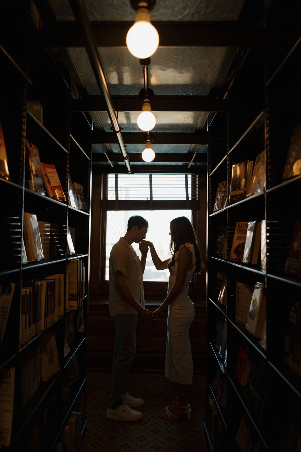 A silhouetted couple shares an intimate moment in a dimly lit library aisle.