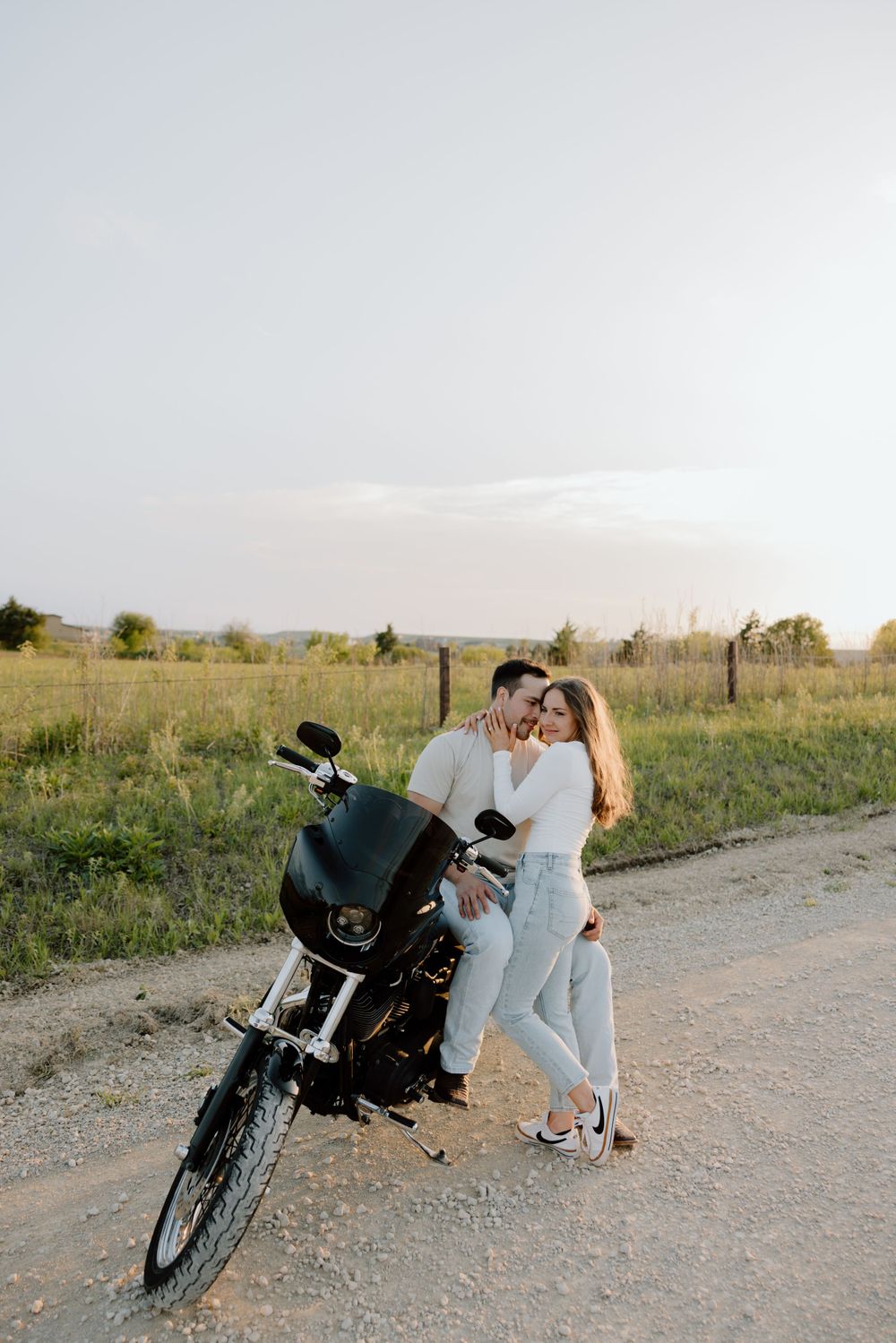 Romantic couple stands beside motorcycle on country road during golden hour.