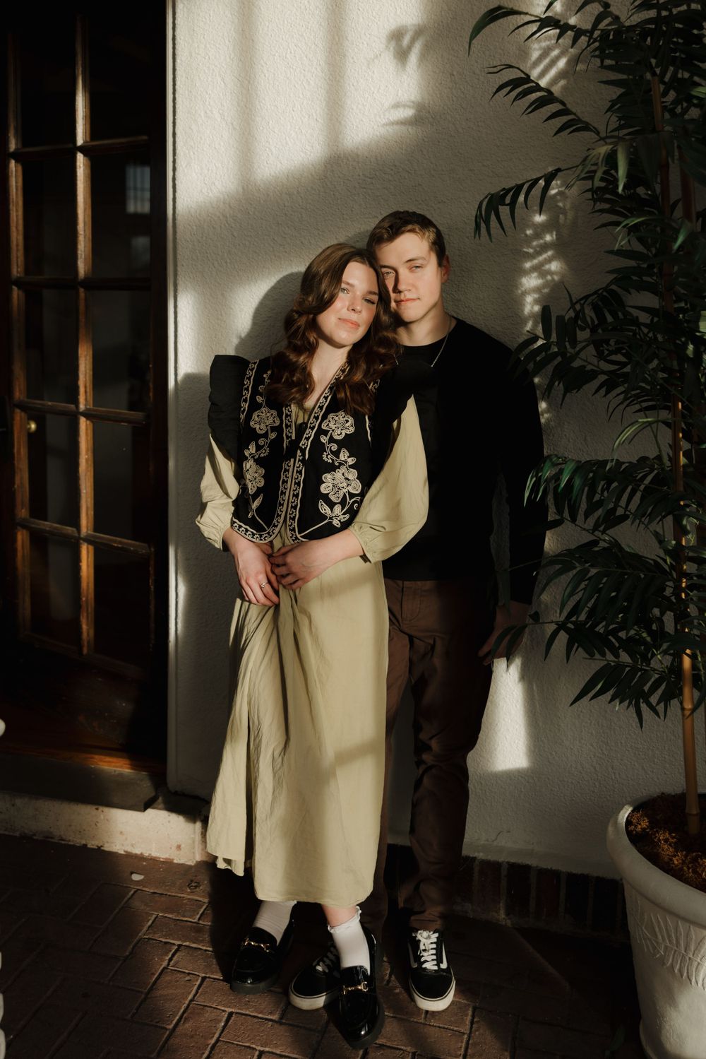 A couple stands in matching dark outfits against a white wall with dramatic shadows and a potted plant beside them.