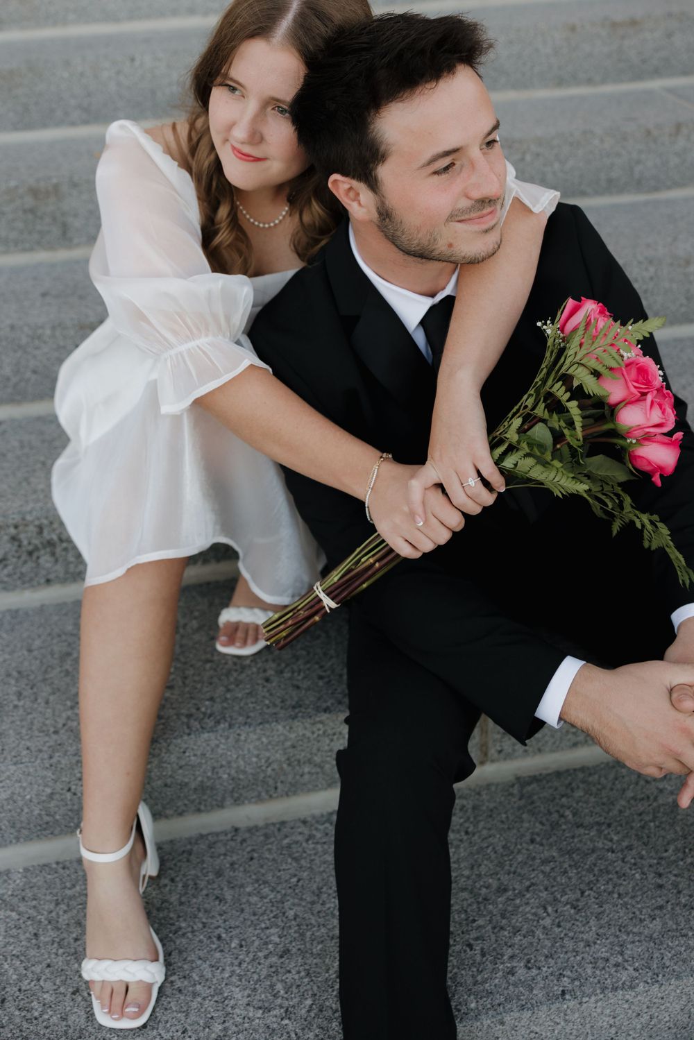 A couple in formal attire shares a tender moment on stone steps with pink roses.