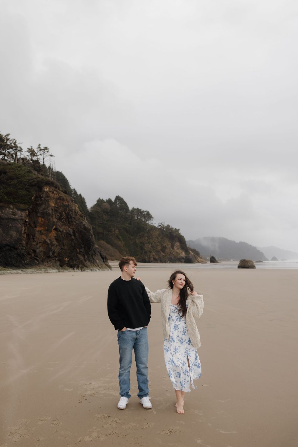 A couple stands together on a misty beach with dramatic coastal cliffs in the background.
