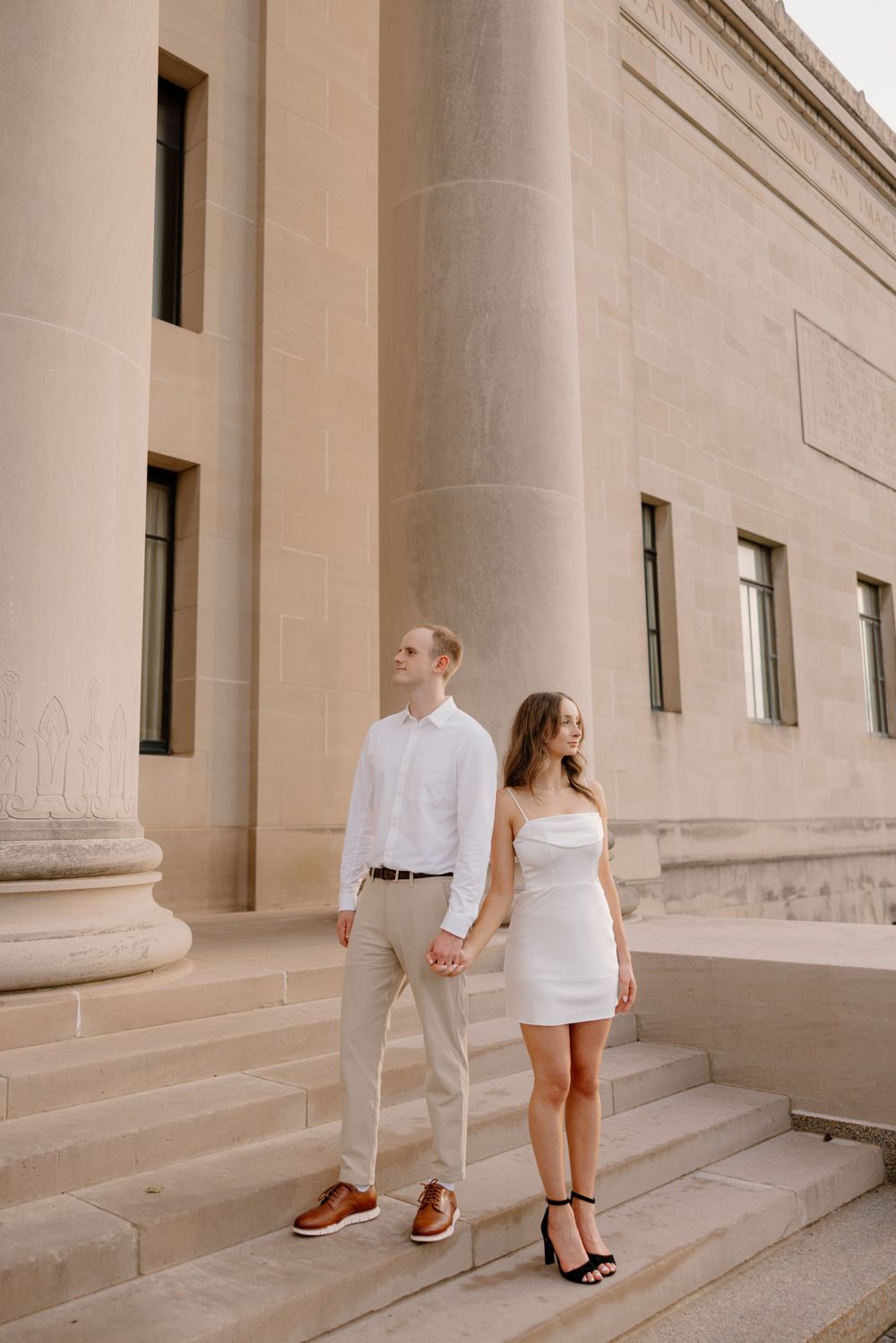 Elegant couple in white attire stand together on grand stone staircase.