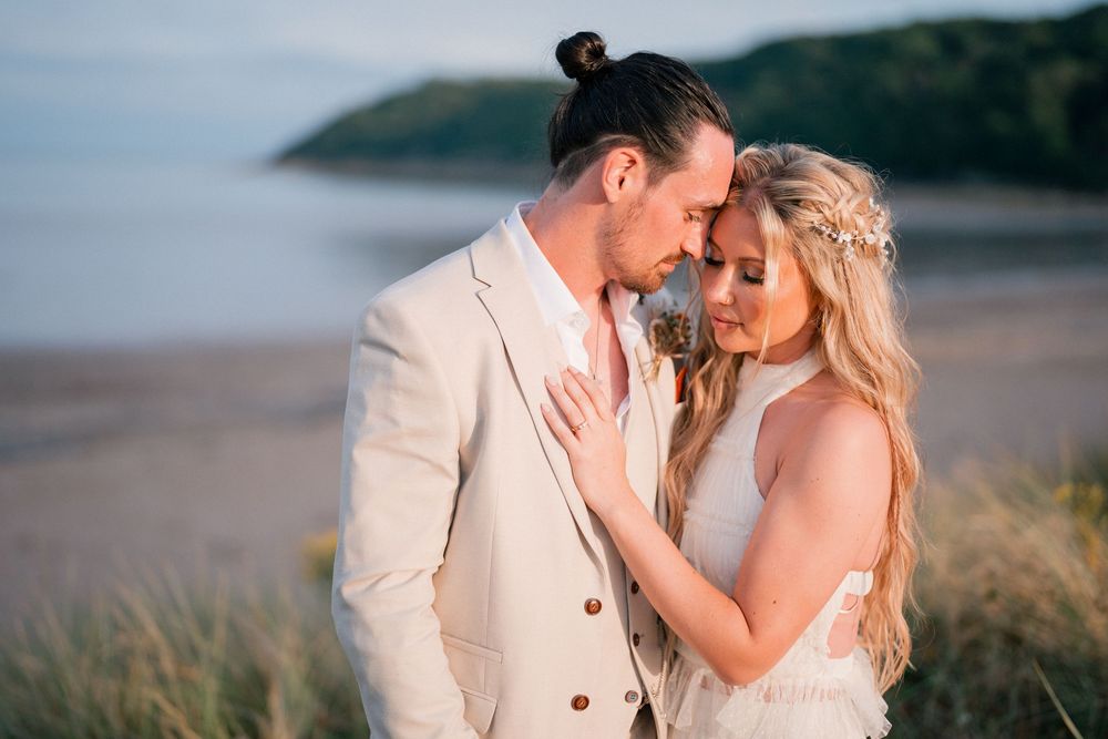 A couple in wedding attire share a romantic moment on a beach, the groom in a beige suit and bride in a white dress.