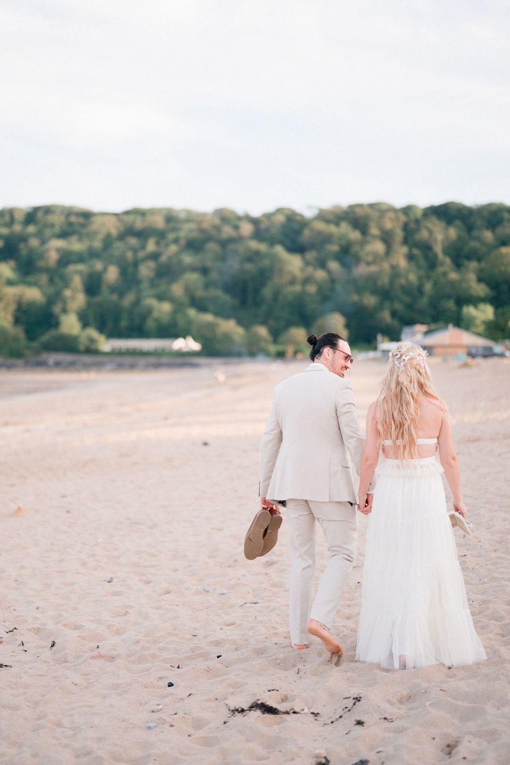 Bride and groom walking hand in hand on sandy beach, groom in beige suit holding hat, bride in white dress.