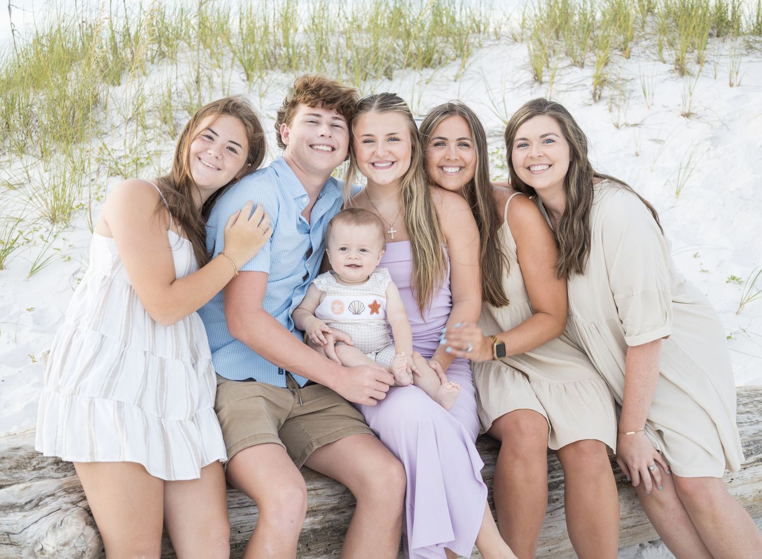 A group sits together on a sandy beach wearing coordinated light colored outfits for a casual photo session.