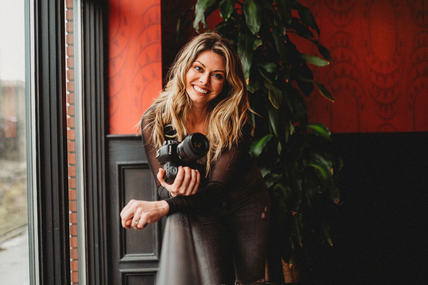 Person in a gray sweater holding a coffee mug stands by a window with a red wall and indoor plant in background. best photographer detroit