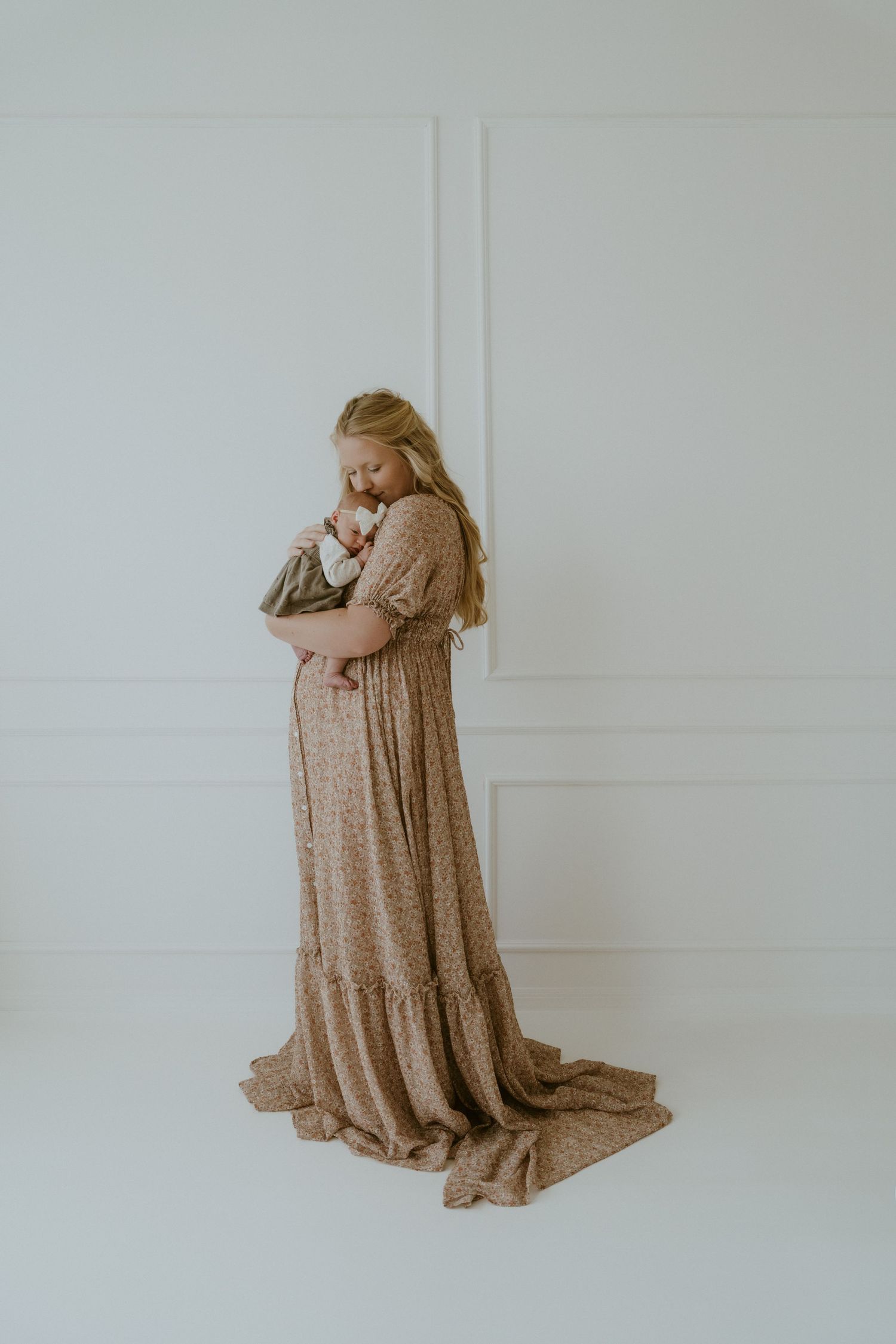 A intimate portrait of mother and baby in a flowing beige dress against white walls.