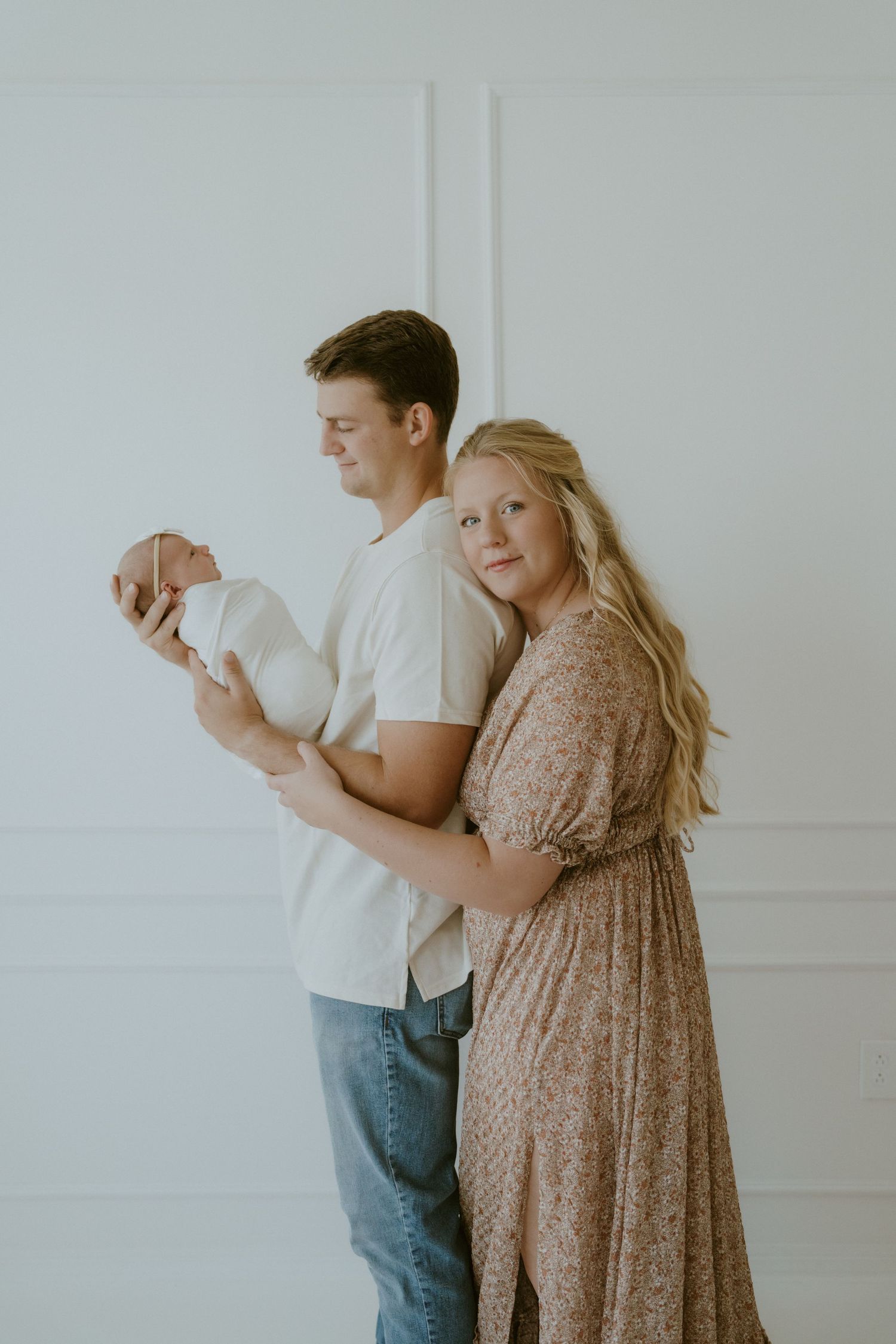 A family portrait with parents holding their newborn in a bright white studio setting.