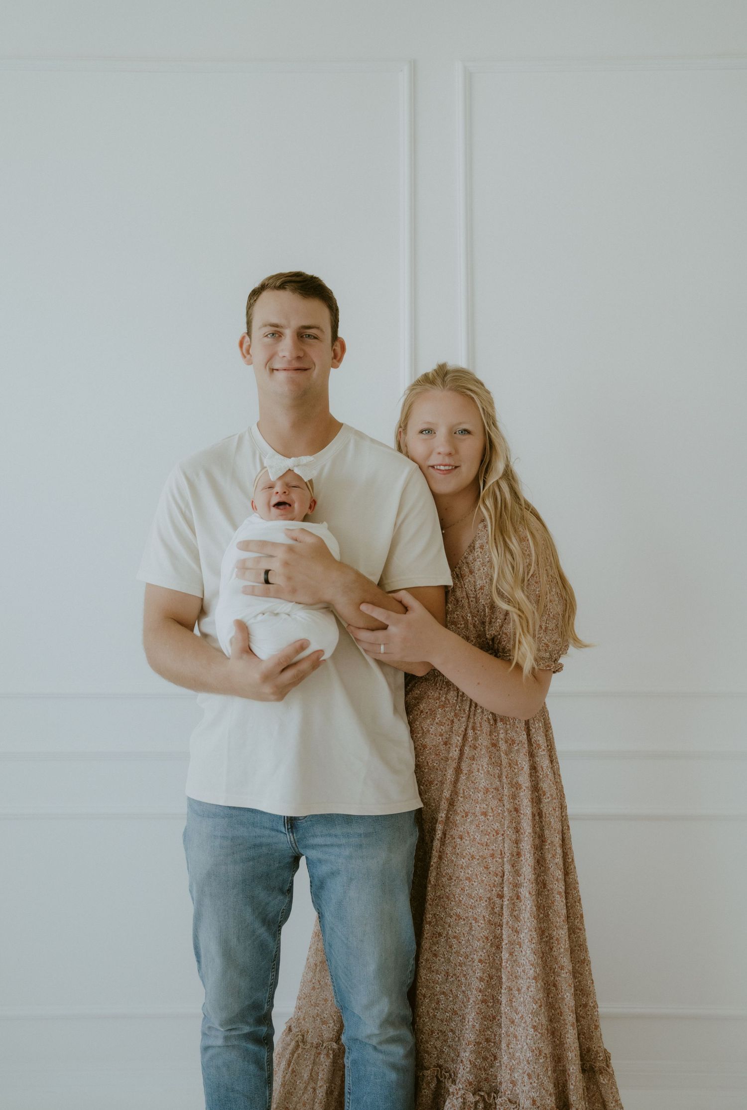 A couple in casual attire holds their newborn baby against a white wall background.