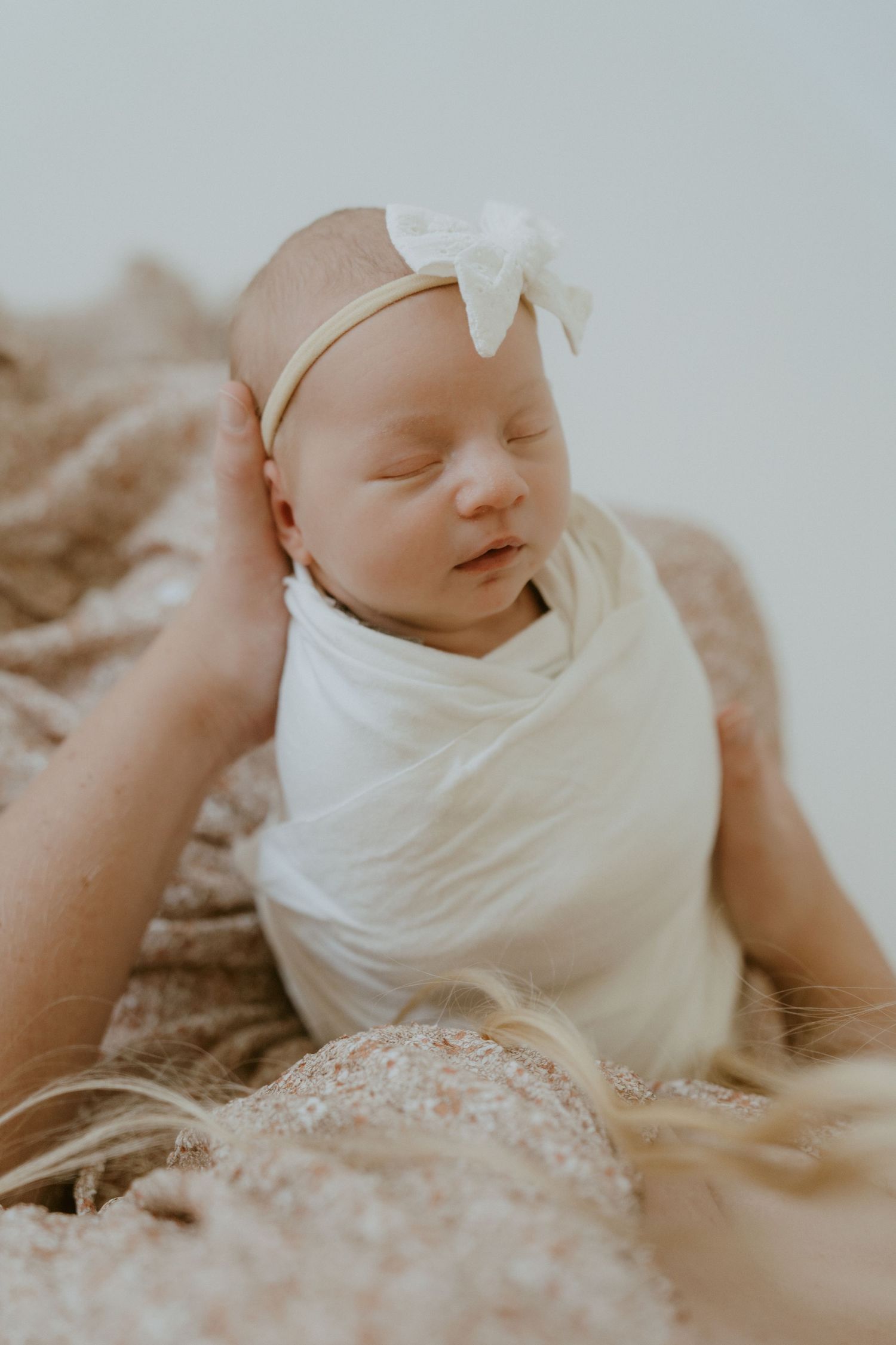 Sleeping newborn baby wrapped in white with a bow headband on textured fabric.