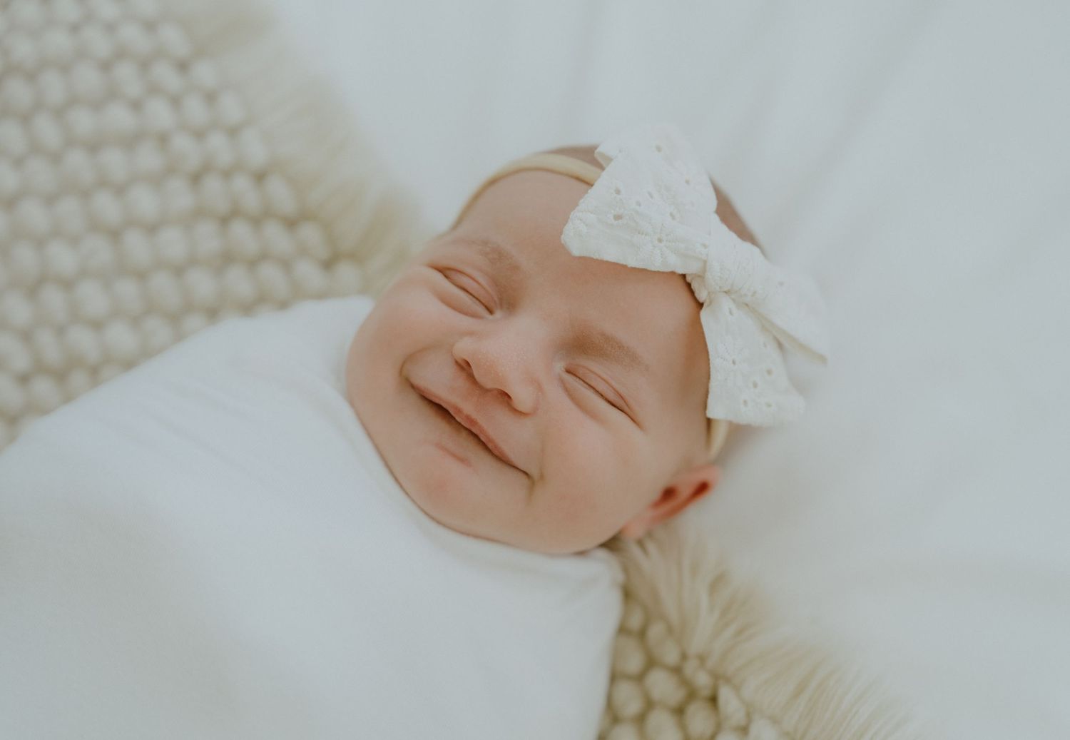 Close up of a smiling newborn baby wearing a white bow headband.