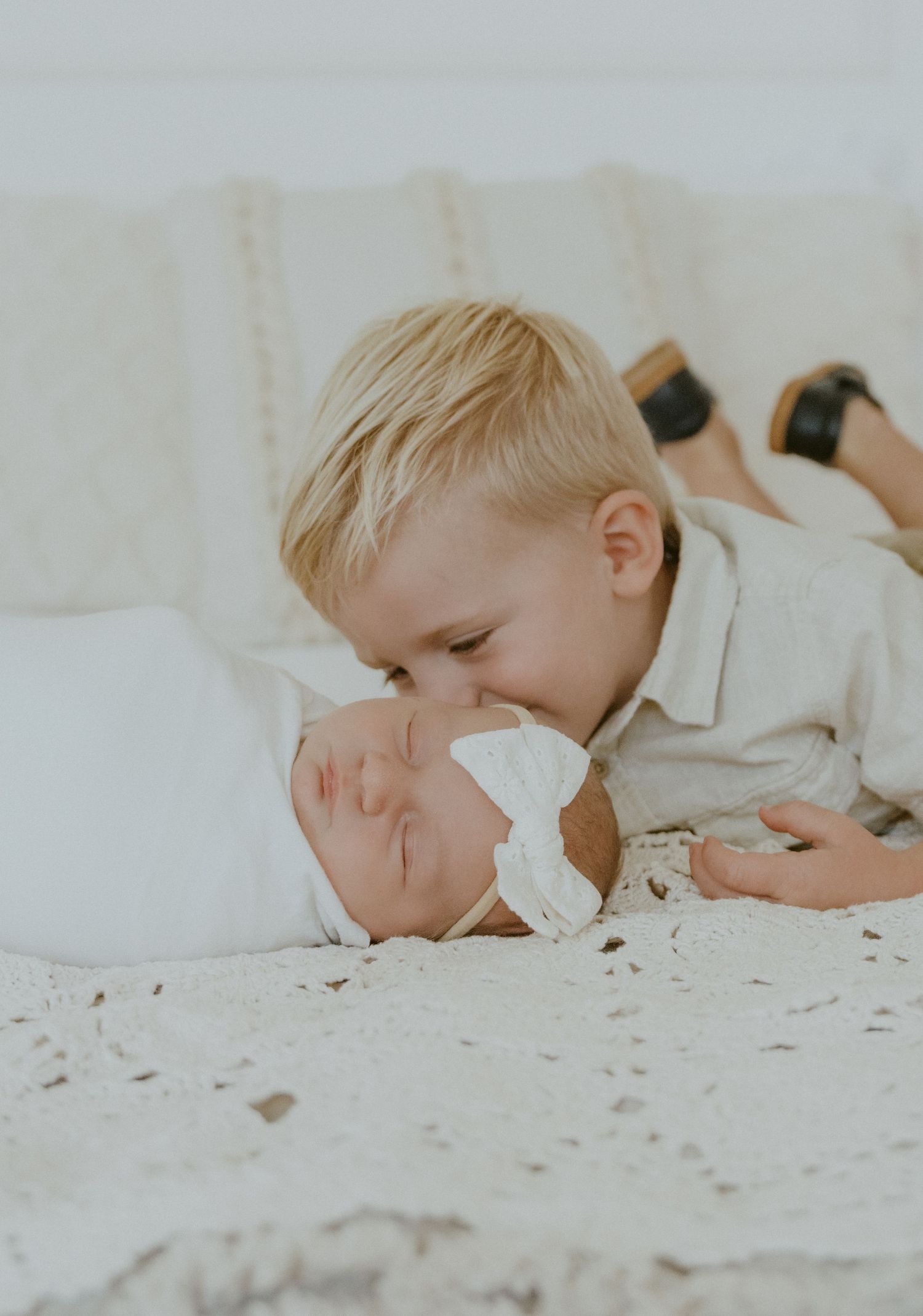 Young child giving gentle kiss to sleeping infant on white bedding.