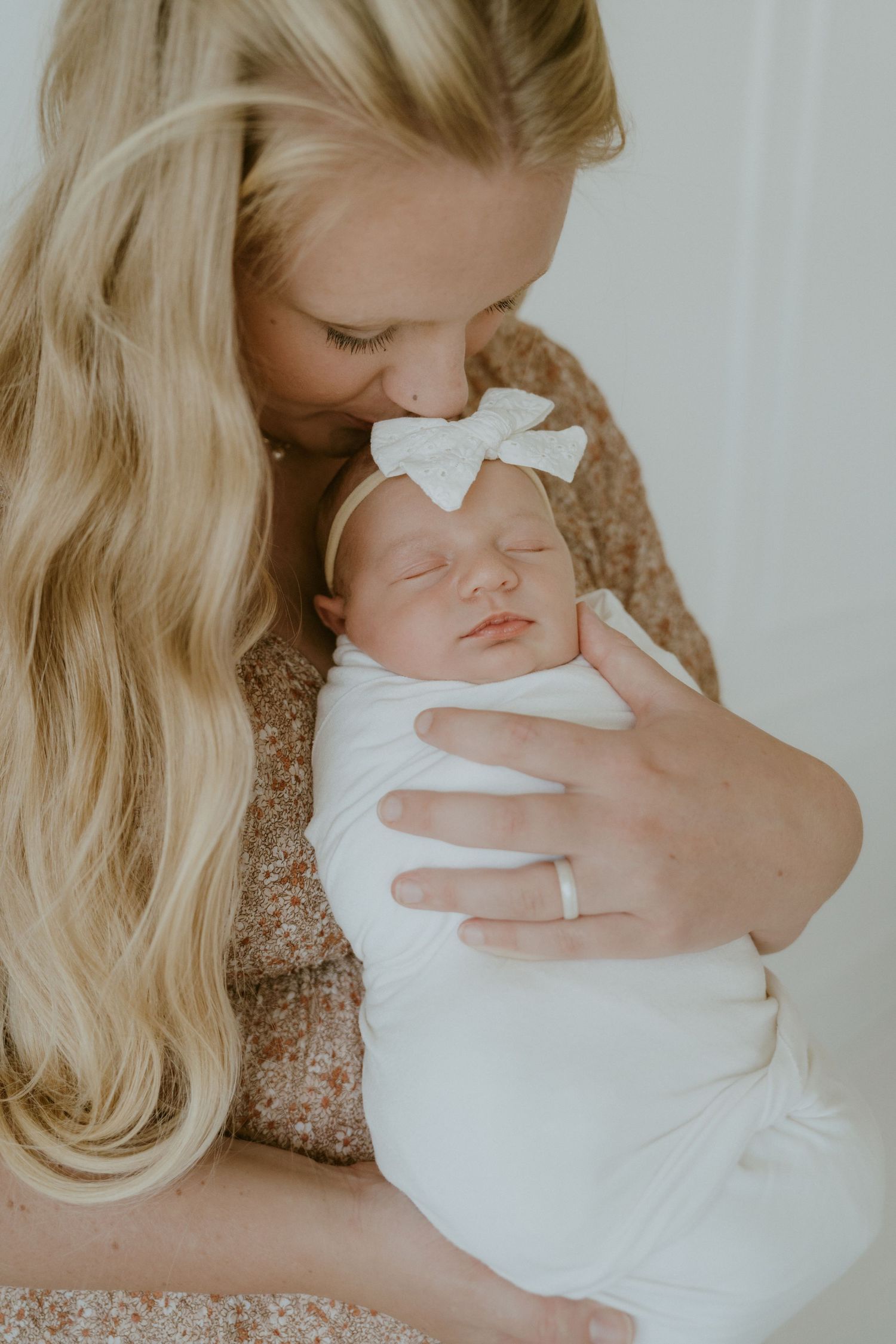 Mother in sparkly dress holding sleeping newborn with white bow.