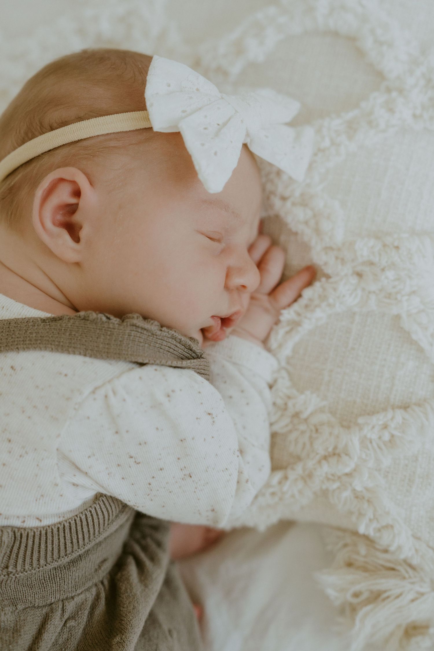 Sleeping newborn baby wearing gray sweater and white bow headband on lacy white bedding.