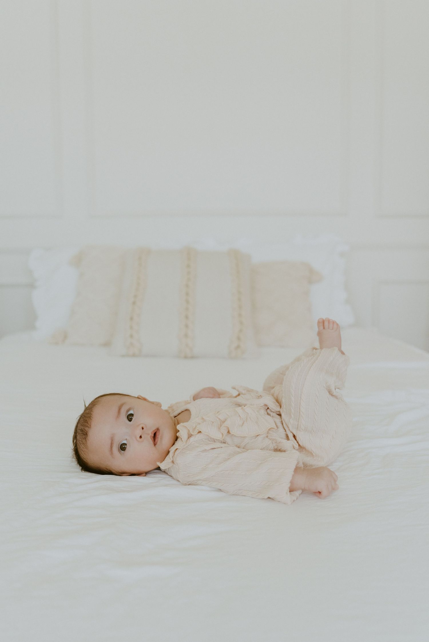 Baby in cream-colored outfit lying on white bedding looking up at camera.