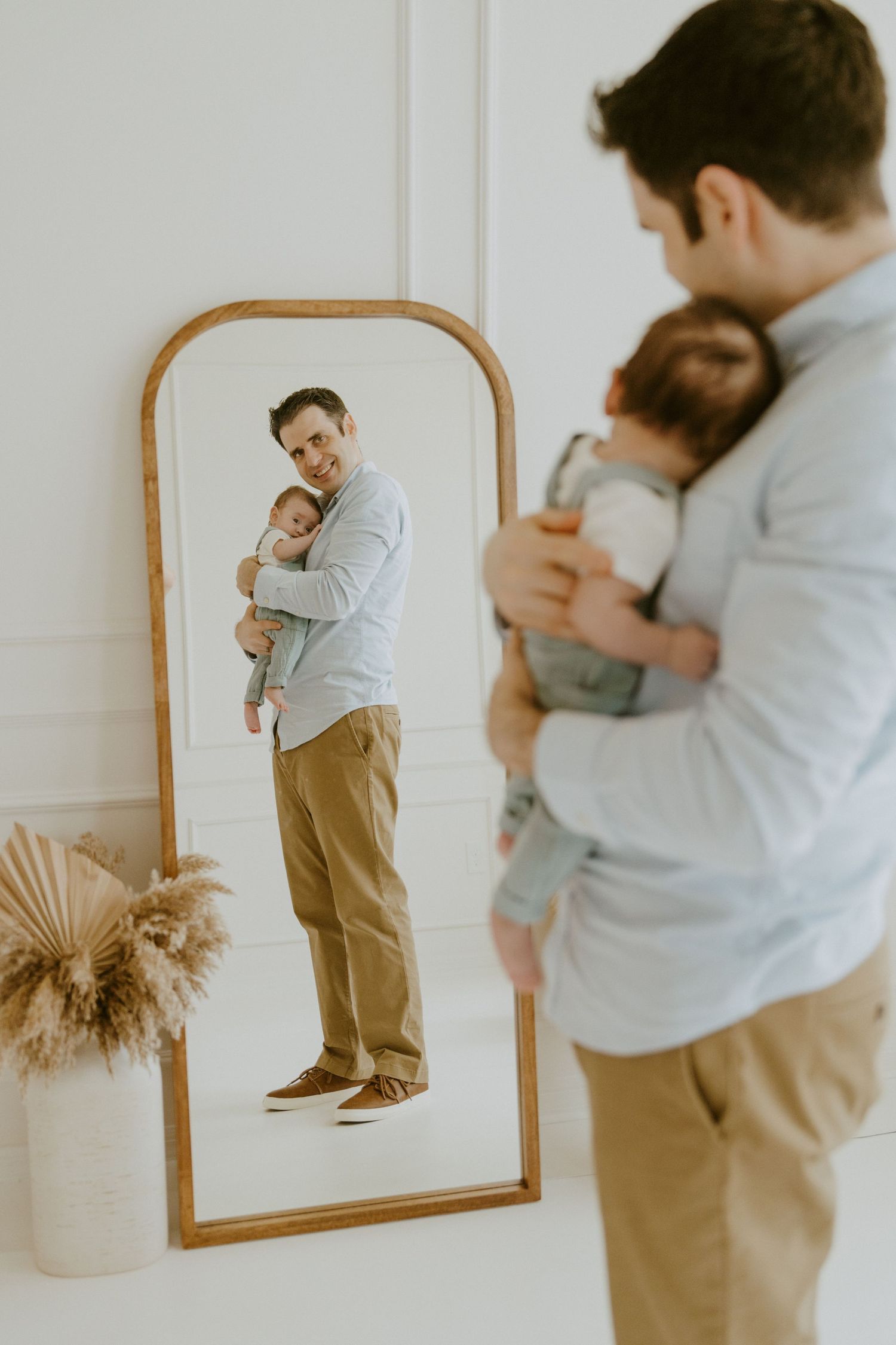 A parent and baby reflected in an arched mirror with pampas grass decor.