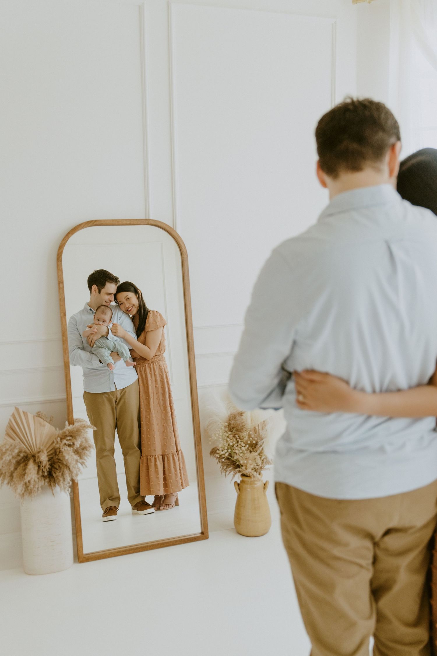 A couple poses in front of a full-length mirror with dried pampas grass decor in a minimalist white room.
