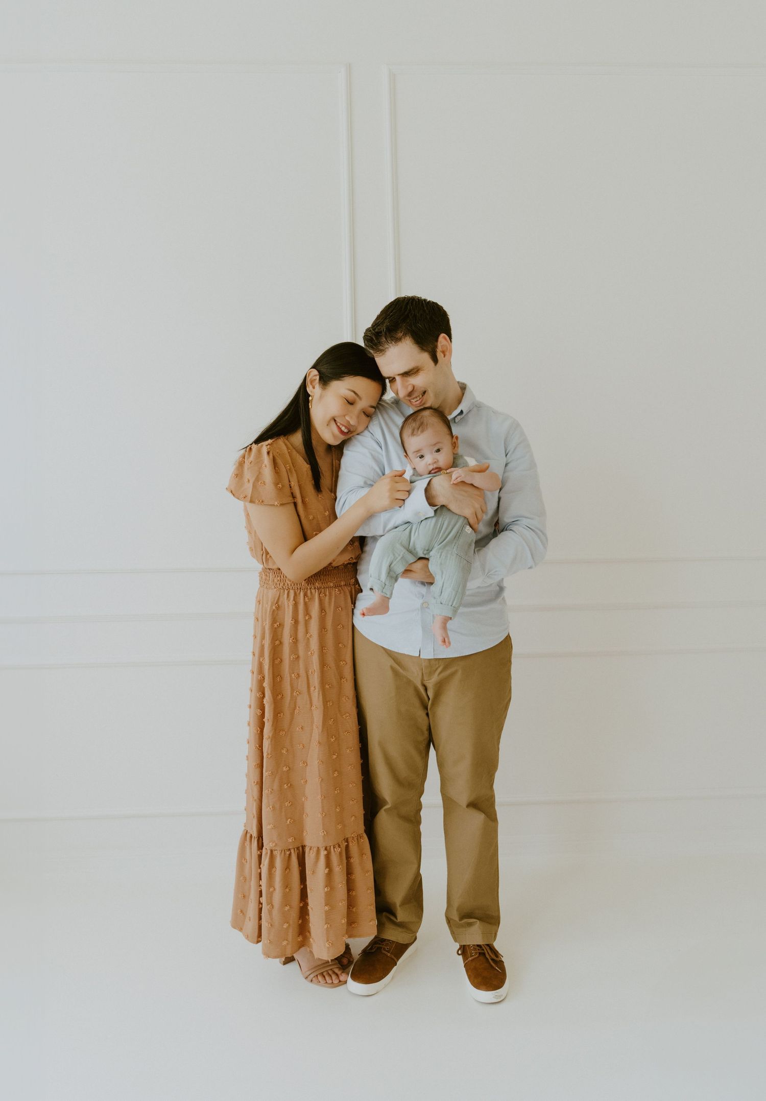A family of three shares an intimate moment together against a white wall background.