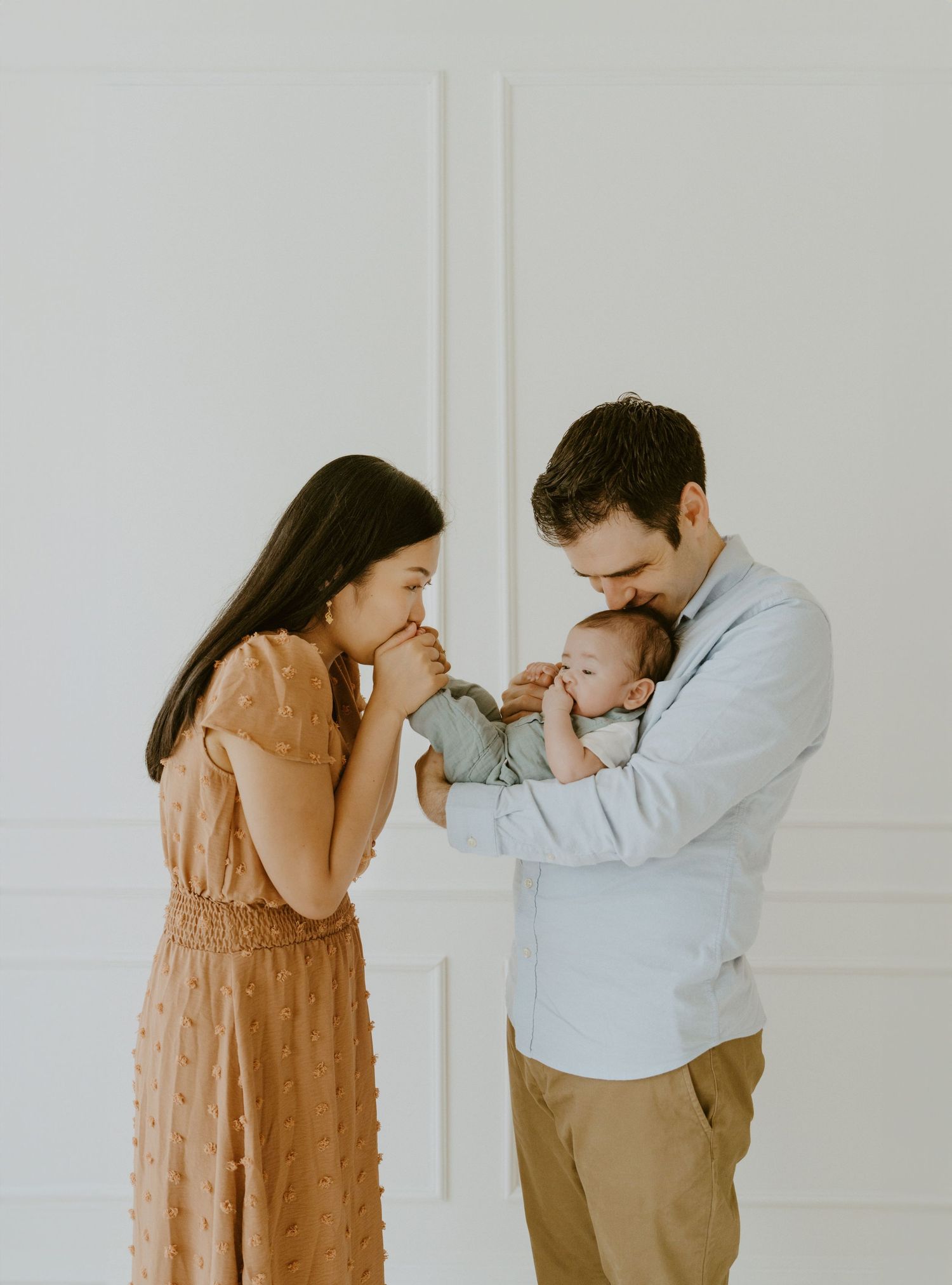 Parents in casual attire share a tender moment with their baby in a white room.
